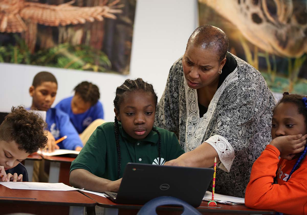 HSA Teacher smiles while kneeling beside a young student in a classroom setting.